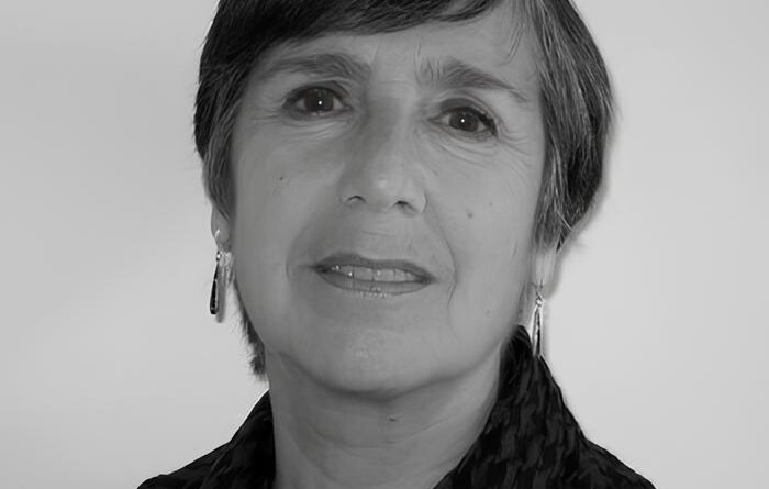 A imagem é um retrato em preto e branco de uma mulher de meia-idade ou mais velha, vista do peito para cima, olhando ligeiramente para a direita da câmera. Ela tem cabelo escuro e curto, com franja. A mulher usa brincos compridos e um lenço ou echarpe com padrão xadrez em tons escuros. Ela tem um sorriso sutil e o fundo da imagem é uma parede clara e uniforme.