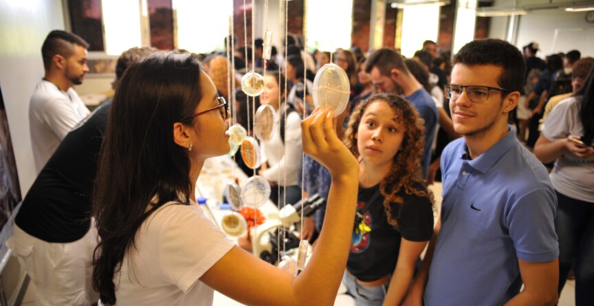 A imagem mostra um grupo de pessoas, principalmente jovens, em um ambiente interno, que parece ser um laboratório ou sala de aula, com iluminação fluorescente no teto. Várias pessoas estão observando e interagindo com uma exposição. Em primeiro plano, uma jovem de cabelos escuros e óculos, vestindo uma camiseta branca, segura uma placa de Petri contra a luz, enquanto explica algo. Uma jovem com cabelos cacheados e um jovem de óculos e camiseta azul, ambos olhando para o objeto, a ouvem atentamente. Ao fundo, mais pessoas estão aglomeradas, observando outras exposições ou conversando.