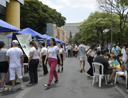 A imagem mostra uma rua movimentada com um evento ao ar livre, possivelmente uma feira ou festival. Várias barracas com toldos azuis e brancos estão alinhadas no lado esquerdo da rua, com pessoas interagindo e passando em frente a elas. No primeiro plano, à esquerda, há um grupo de pessoas em torno de uma das barracas. Uma mulher de calças cor-de-rosa e blusa branca caminha pelo centro da rua. Mais à direita, outro grupo de pessoas está sentado em cadeiras de plástico brancas em torno de uma mesa, com crianças e adultos presentes. Ao fundo, árvores e edifícios compõem a paisagem urbana. O céu é claro, indicando um dia ensolarado.