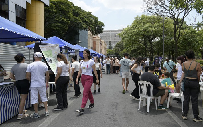 A imagem mostra uma rua movimentada com um evento ao ar livre, possivelmente uma feira ou festival. Várias barracas com toldos azuis e brancos estão alinhadas no lado esquerdo da rua, com pessoas interagindo e passando em frente a elas. No primeiro plano, à esquerda, há um grupo de pessoas em torno de uma das barracas. Uma mulher de calças cor-de-rosa e blusa branca caminha pelo centro da rua. Mais à direita, outro grupo de pessoas está sentado em cadeiras de plástico brancas em torno de uma mesa, com crianças e adultos presentes. Ao fundo, árvores e edifícios compõem a paisagem urbana. O céu é claro, indicando um dia ensolarado.