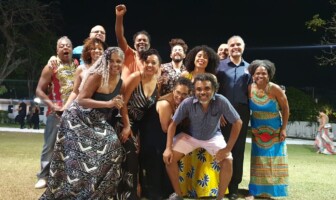 A imagem mostra um grande grupo de pessoas, de diversas idades e etnias, posando para uma foto em um gramado à noite. Eles estão sorrindo e em poses descontraídas, com alguns abraçados. Um homem no centro do grupo levanta o punho. As pessoas vestem roupas casuais e coloridas, com alguns trajes apresentando estampas vibrantes. O fundo da imagem é um gramado escuro, com árvores e algumas luzes fracas visíveis ao longe, indicando um ambiente externo.