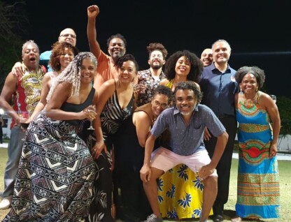 A imagem mostra um grande grupo de pessoas, de diversas idades e etnias, posando para uma foto em um gramado à noite. Eles estão sorrindo e em poses descontraídas, com alguns abraçados. Um homem no centro do grupo levanta o punho. As pessoas vestem roupas casuais e coloridas, com alguns trajes apresentando estampas vibrantes. O fundo da imagem é um gramado escuro, com árvores e algumas luzes fracas visíveis ao longe, indicando um ambiente externo.