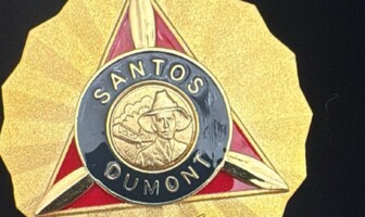 A imagem é um close-up de uma medalha dourada com formato de sol ou estrela. No centro da medalha, há um triângulo invertido em vermelho, emoldurado por uma borda dourada. Dentro do triângulo, há um círculo preto com as palavras "SANTOS DUMONT" em letras douradas. No centro do círculo, um busto de um homem com chapéu e um avião são representados em dourado. Abaixo do triângulo e do círculo, um emblema alado é visível. A medalha tem um anel dourado na parte superior para ser pendurada, e o fundo da imagem é preto.