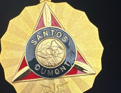 A imagem é um close-up de uma medalha dourada com formato de sol ou estrela. No centro da medalha, há um triângulo invertido em vermelho, emoldurado por uma borda dourada. Dentro do triângulo, há um círculo preto com as palavras "SANTOS DUMONT" em letras douradas. No centro do círculo, um busto de um homem com chapéu e um avião são representados em dourado. Abaixo do triângulo e do círculo, um emblema alado é visível. A medalha tem um anel dourado na parte superior para ser pendurada, e o fundo da imagem é preto.