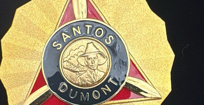A imagem é um close-up de uma medalha dourada com formato de sol ou estrela. No centro da medalha, há um triângulo invertido em vermelho, emoldurado por uma borda dourada. Dentro do triângulo, há um círculo preto com as palavras "SANTOS DUMONT" em letras douradas. No centro do círculo, um busto de um homem com chapéu e um avião são representados em dourado. Abaixo do triângulo e do círculo, um emblema alado é visível. A medalha tem um anel dourado na parte superior para ser pendurada, e o fundo da imagem é preto.