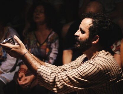 A imagem mostra um homem de pele escura, com barba e cabelos escuros e encaracolados, ajoelhado e olhando para o lado direito da foto. Ele veste uma camisa listrada em tons de bege e marrom. O homem está com os braços estendidos, segurando uma pequena tigela metálica prateada com as duas mãos. A iluminação é dramática, com um feixe de luz destacando o homem e a tigela contra um fundo escuro e desfocado, onde outras pessoas são visíveis.