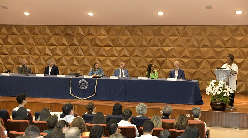 A imagem mostra um auditório com um palco elevado de madeira. No palco, uma longa mesa coberta por uma toalha azul escura, com um emblema dourado na frente, acomoda sete pessoas sentadas, todas vestidas com trajes formais. À direita do palco, uma mulher de pele escura e cabelos crespos, vestindo uma blusa clara, está em pé em um púlpito, falando em um microfone. Um arranjo floral branco está ao lado do púlpito. O fundo do palco é uma parede de madeira com um padrão geométrico tridimensional. Em primeiro plano, o público está sentado em cadeiras estofadas em tons de marrom.