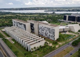 A imagem é uma vista aérea de um complexo arquitetônico moderno, que inclui vários edifícios de cor clara com detalhes escuros, em um ambiente paisagístico com gramados verdes e algumas árvores, sob um céu parcialmente nublado. No centro da imagem, um longo edifício horizontal com um padrão quadriculado em suas janelas se estende. Em frente a ele, uma série de estruturas menores e uniformes, com telhados em ângulo, formam um design em cascata. Ao fundo, um grande lago é visível, com a paisagem natural e urbana do outro lado. Ruas e veículos são visíveis nas bordas da imagem.