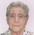 A imagem é um retrato de uma mulher idosa, vista do peito para cima, olhando diretamente para a câmera. Ela tem cabelo grisalho penteado para trás e usa óculos de aros claros. A mulher veste uma blusa clara com um padrão floral em tons de azul e verde. Ela também usa um colar discreto de pérolas. O fundo da imagem é claro e uniforme.