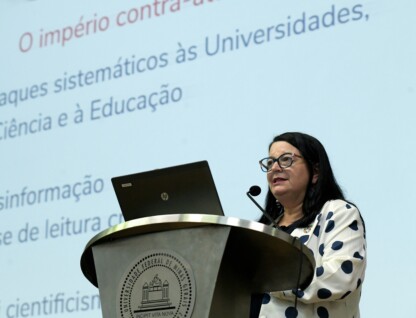 A imagem mostra uma mulher de cabelos escuros e lisos, usando óculos de armação colorida, em pé atrás de um púlpito. Ela veste um blazer branco com bolinhas azuis e um broche em sua lapela. A mulher está falando em um microfone no púlpito, que possui um emblema em relevo. Um laptop aberto é visível sobre o púlpito. Ao fundo, uma tela de projeção branca exibe texto em letras azuis e vermelhas, com frases como "Ataques sistemáticos às Universidades" e "Desinformação".