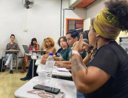 A imagem mostra um grupo de pessoas sentadas em carteiras escolares, em uma sala de aula ou workshop. As pessoas, em sua maioria jovens, estão atentas e algumas estão com notebooks abertos ou fazendo anotações. Em primeiro plano, à direita, uma mulher de pele escura com cabelo crespo e um turbante amarelo e rosa está falando, com um microfone na mão e gesticulando. Ela está de costas para a câmera. Ao longo das fileiras de carteiras, outras pessoas são visíveis, com diferentes tons de pele e estilos. Um ventilador está fixado na parede branca ao fundo.
