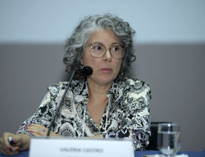 A imagem mostra uma mulher de meia-idade com cabelos grisalhos e cacheados, usando óculos de armação clara. Ela está sentada a uma mesa coberta por uma toalha azul escura, com um microfone preto à sua frente. A mulher veste uma blusa estampada em preto e branco com um padrão paisley. Suas mãos estão apoiadas na mesa, e ela segura uma caneta na mão direita. Um copo com água é visível à sua direita. O fundo da imagem é uma parede lisa de cor clara, com uma faixa escura na parte inferior.