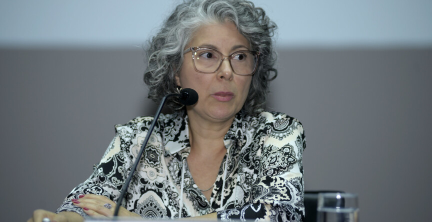 A imagem mostra uma mulher de meia-idade com cabelos grisalhos e cacheados, usando óculos de armação clara. Ela está sentada a uma mesa coberta por uma toalha azul escura, com um microfone preto à sua frente. A mulher veste uma blusa estampada em preto e branco com um padrão paisley. Suas mãos estão apoiadas na mesa, e ela segura uma caneta na mão direita. Um copo com água é visível à sua direita. O fundo da imagem é uma parede lisa de cor clara, com uma faixa escura na parte inferior.