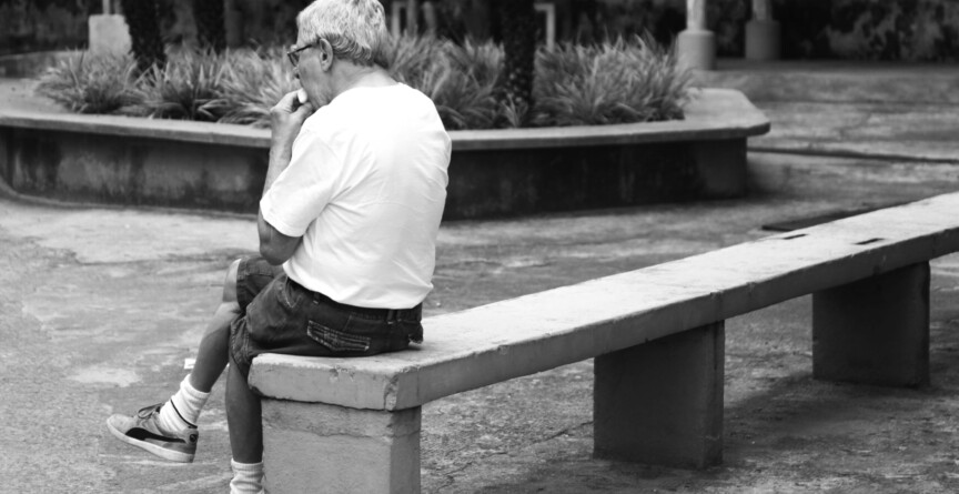 Homem idoso de camisa branco sentado em um banco de praça pública