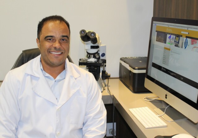 Professor Adriano de Paula Sabino veste um jaleco branco e está sentado em frente a um computador