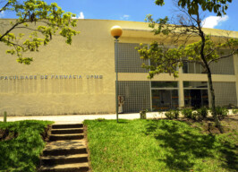 A imagem mostra a fachada de um edifício institucional de cor bege claro, que abriga a "FACULDADE DE FARMÁCIA UFMG", conforme indicado por uma inscrição em relevo na parede esquerda. Duas árvores com folhagem verde emolduram a cena, uma de cada lado. Um poste de iluminação com uma luminária esférica amarela está posicionado na frente do prédio.