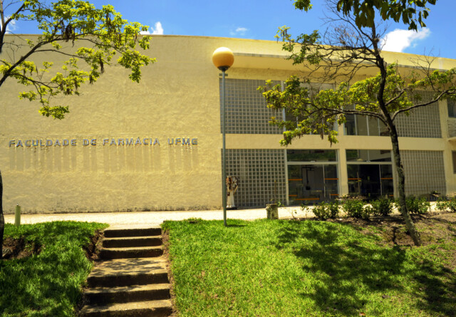 A imagem mostra a fachada de um edifício institucional de cor bege claro, que abriga a "FACULDADE DE FARMÁCIA UFMG", conforme indicado por uma inscrição em relevo na parede esquerda. Duas árvores com folhagem verde emolduram a cena, uma de cada lado. Um poste de iluminação com uma luminária esférica amarela está posicionado na frente do prédio.