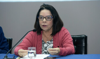 Denise Pires de Carvalho é uma mulher de cabelo preto, que usa óculos e veste uma blusa vermelho. Está sentada diante de uma mesa com forro azul; sobre a mesa estão um copo d´água e papéis
