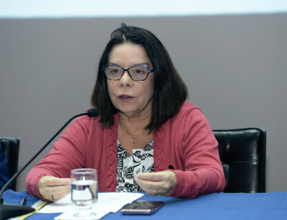 Denise Pires de Carvalho é uma mulher de cabelo preto, que usa óculos e veste uma blusa vermelho. Está sentada diante de uma mesa com forro azul; sobre a mesa estão um copo d´água e papéis