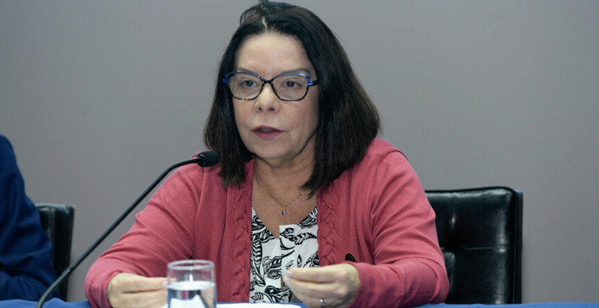 Denise Pires de Carvalho é uma mulher de cabelo preto, que usa óculos e veste uma blusa vermelho. Está sentada diante de uma mesa com forro azul; sobre a mesa estão um copo d´água e papéis