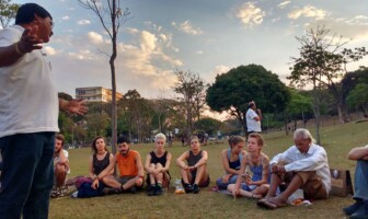 Um professor vestido de camisa branca conversa com um grupo de alunos sentado em um gramado no campus Pampulha