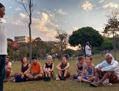 Um professor vestido de camisa branca conversa com um grupo de alunos sentado em um gramado no campus Pampulha