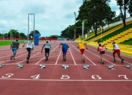 Grupo de atletas correm em uma pista avermelhada com raias numeradas (4, 5 e 6 são os números visíveis): à direita, vê-se uma arquibancada em tom amarelo