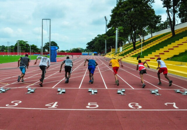 Grupo de atletas correm em uma pista avermelhada com raias numeradas (4, 5 e 6 são os números visíveis): à direita, vê-se uma arquibancada em tom amarelo