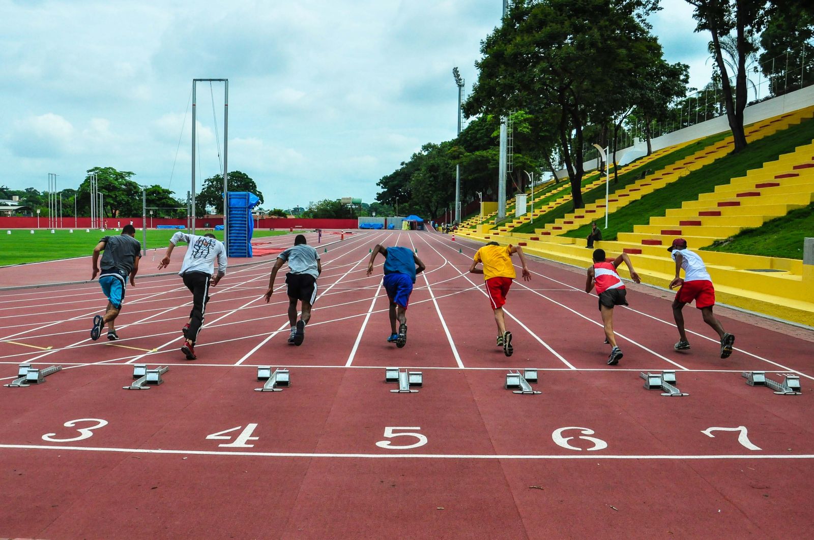 Grupo de atletas correm em uma pista avermelhada com raias numeradas (4, 5 e 6 são os números visíveis): à direita, vê-se uma arquibancada em tom amarelo