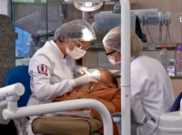 Profissionais atendem paciente em clínica da Faculdade de Odontogia. Elas usam jalecos brancos, toucas na cabeça e máscaras de proteção no rosto.