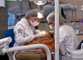 Profissionais atendem paciente em clínica da Faculdade de Odontogia. Elas usam jalecos brancos, toucas na cabeça e máscaras de proteção no rosto.