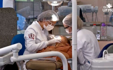 Profissionais atendem paciente em clínica da Faculdade de Odontogia. Elas usam jalecos brancos, toucas na cabeça e máscaras de proteção no rosto.