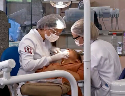 Profissionais atendem paciente em clínica da Faculdade de Odontogia. Elas usam jalecos brancos, toucas na cabeça e máscaras de proteção no rosto.