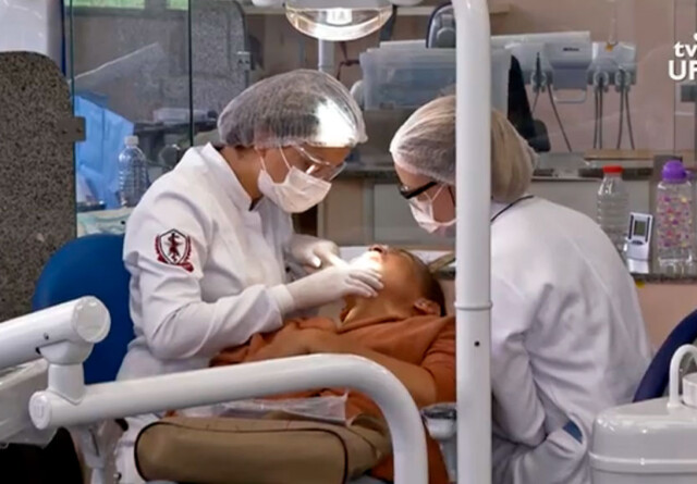 Profissionais atendem paciente em clínica da Faculdade de Odontogia. Elas usam jalecos brancos, toucas na cabeça e máscaras de proteção no rosto.
