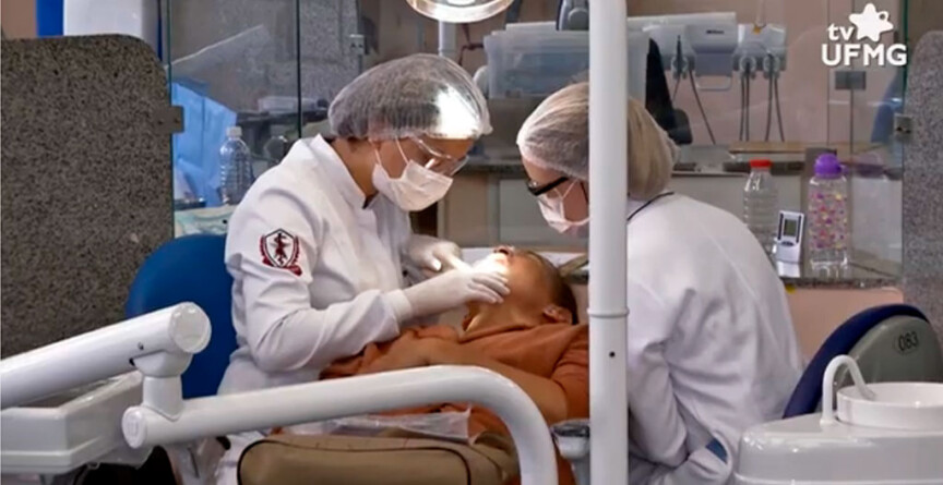 Profissionais atendem paciente em clínica da Faculdade de Odontogia. Elas usam jalecos brancos, toucas na cabeça e máscaras de proteção no rosto.