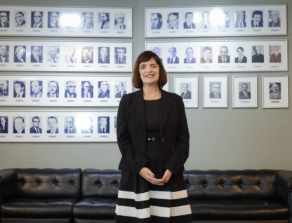 Mulher de cabelo escuro, roupa preta, com detalhes em branco posa diante de um quadro com personalidades ao fundo