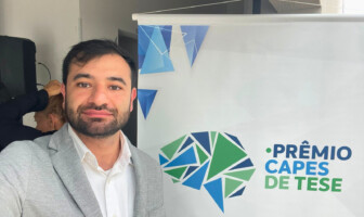 Imagem mostra homem de cabelo preto vestido com paletó cinza e camisa branca à frente de um banner em que se lê "Prêmio Capes de Teses"