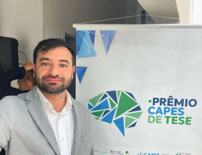 Imagem mostra homem de cabelo preto vestido com paletó cinza e camisa branca à frente de um banner em que se lê "Prêmio Capes de Teses"