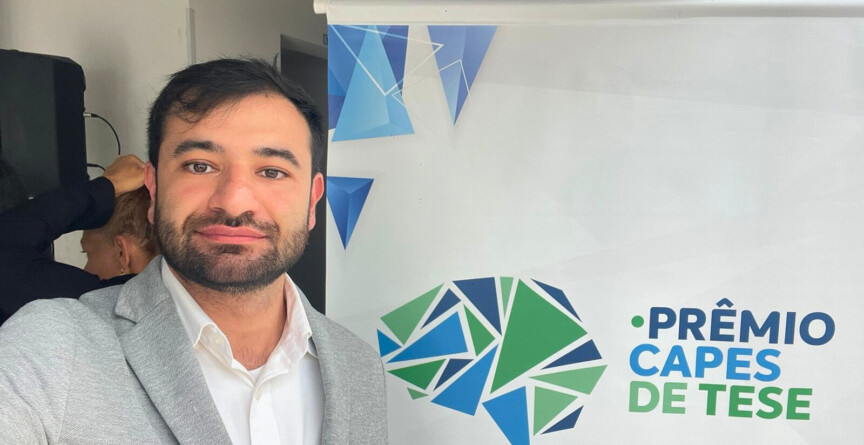 Imagem mostra homem de cabelo preto vestido com paletó cinza e camisa branca à frente de um banner em que se lê "Prêmio Capes de Teses"