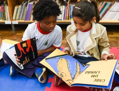 Duas crianças (um garoto e uma garota) estão sentados em um piso de uma biblioteca com livros infantis no colo
