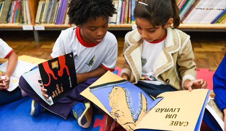 Duas crianças (um garoto e uma garota) estão sentados em um piso de uma biblioteca com livros infantis no colo