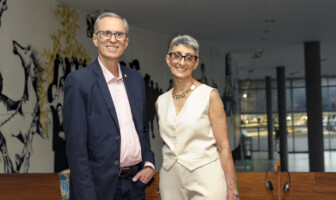 À esquerda, o novo reitor Alessandro Fernandes Moreira. Ele usa óculos e veste um paletó azul. Ao seu lado, a nova vice-reitora Alamanda Kfoury. Ela veste um conjunto creme e usa óculos. Eles estão no mezanino do prédio da Reitoria, tendo ao fundo um painel artístico