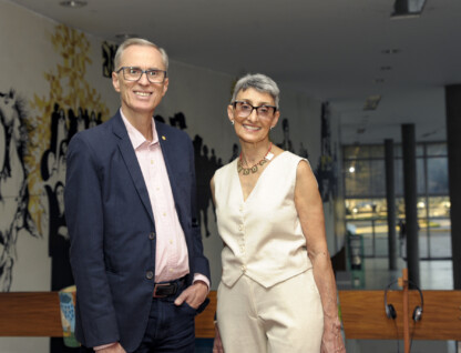 À esquerda, o novo reitor Alessandro Fernandes Moreira. Ele usa óculos e veste um paletó azul. Ao seu lado, a nova vice-reitora Alamanda Kfoury. Ela veste um conjunto creme e usa óculos. Eles estão no mezanino do prédio da Reitoria, tendo ao fundo um painel artístico