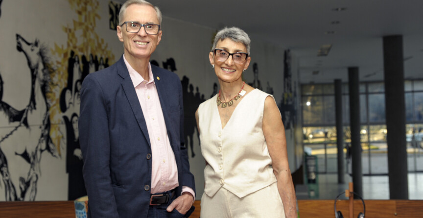 À esquerda, o novo reitor Alessandro Fernandes Moreira. Ele usa óculos e veste um paletó azul. Ao seu lado, a nova vice-reitora Alamanda Kfoury. Ela veste um conjunto creme e usa óculos. Eles estão no mezanino do prédio da Reitoria, tendo ao fundo um painel artístico