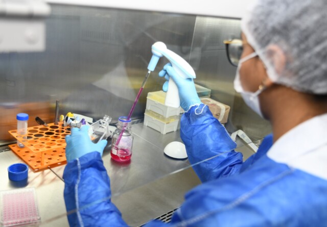 A pesquisadora Nathalia veste roupa de proteção azul enquanto administra material dentro do laboratório