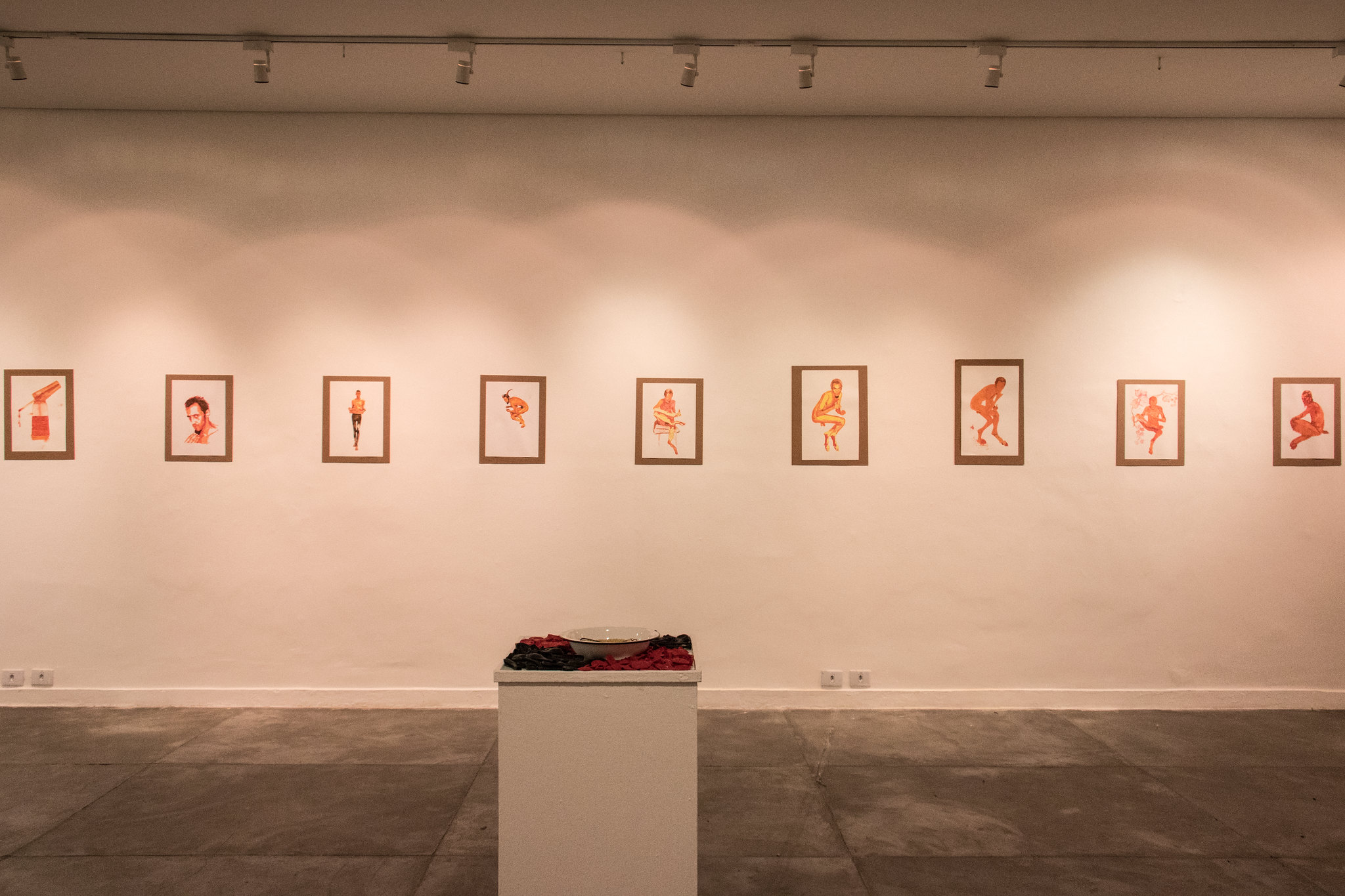 Quadros pendurados na parede, compondo a exposição BooM de Remanery, que ocorreu no Centro Cultural UFMG. Os quadros estão na vertical e contém pinturas de corpos variando entre posições agachados, sentados ou em pé.