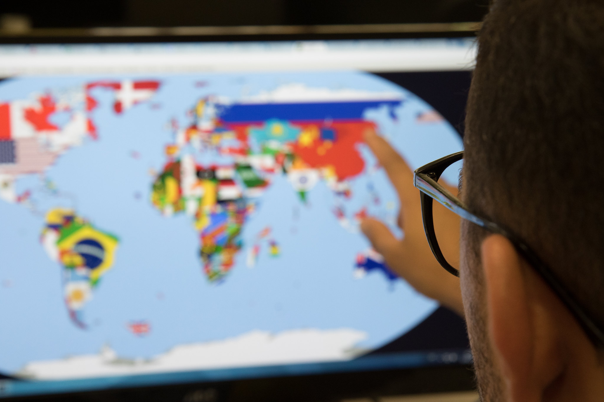 Desfocado, ao fundo, um mapa mundi na tela de um computador. Os países estão delimitados por suas respectivas bandeiras.