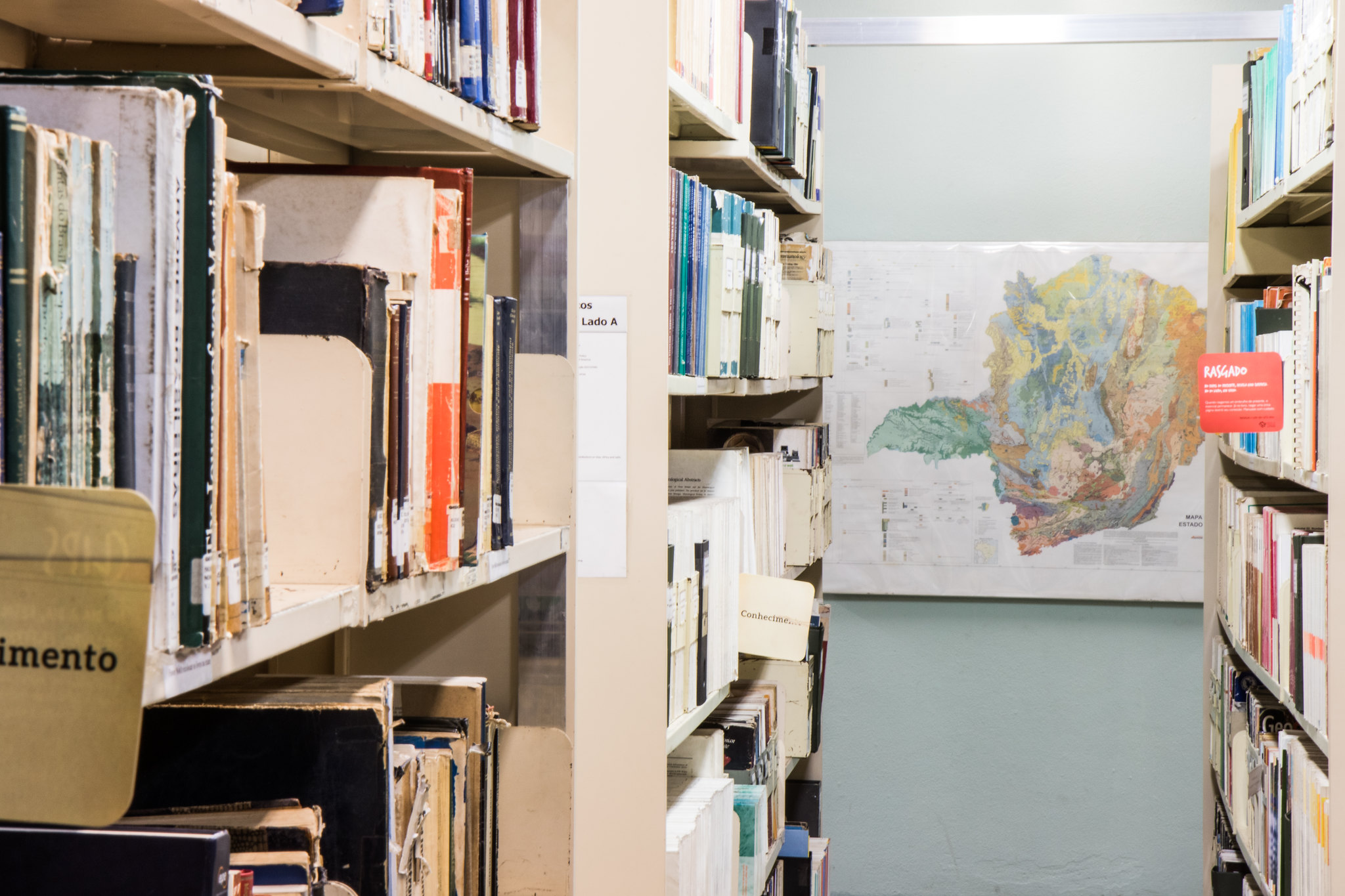 Corredor entre estantes de metal bege com livros e publicações antigas em uma biblioteca. Ao fundo, em uma parede clara, há um grande mapa geológico colorido do estado de Minas Gerais.