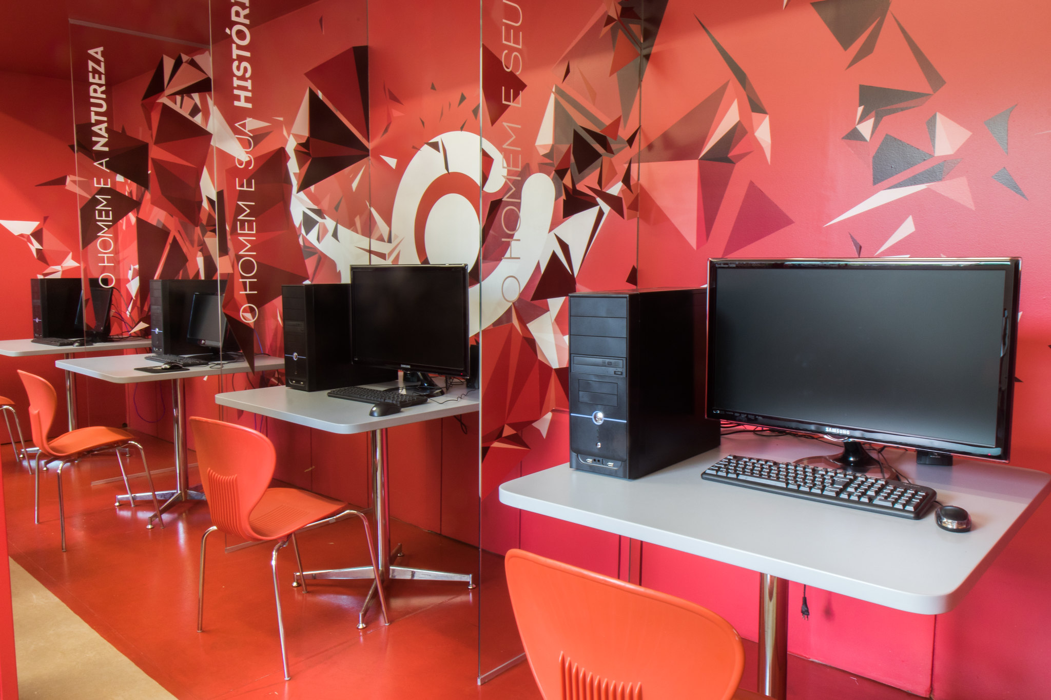 Estações de trabalho compostas por mesas cinzas, computadores de gabinete preto, monitores, teclados, mouses e cadeiras alaranjadas disponíveis no Museu de História Natural. A parede ao fundo e o chão são vermelhos, com grafismos geométricos e frases como "O homem e sua história" e "O homem e sua natureza".