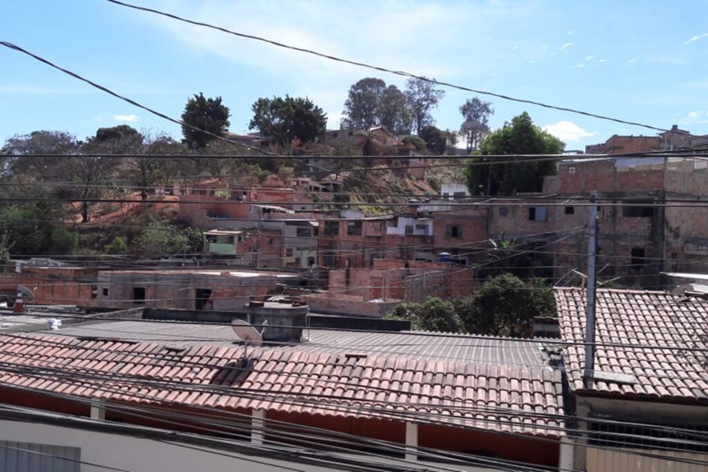 A imagem é a frente de um cartão postal, apresentando uma fotografia em que predominam tons terrosos. O dia está claro. Trata-se de um pequeno aglomerado de casas em terreno íngreme. Em primeiro plano, muitos fios de energia atravessam a imagem em sentido horizontal. Em segundo plano, na faixa inferior, avista-se de cima alguns telhados, o mais extenso deles com uma caixa d’água e uma antena de televisão. Ao fundo, na faixa central, um morro em que, do lado direito, predominam casas de tijolo aparente, algumas ainda em construção, e do lado esquerdo e no topo predominam árvores, algumas delas secas. Na faixa superior, o céu está azul com poucas nuvens brancas. #PraCegoVer #ParaTodosVerem 